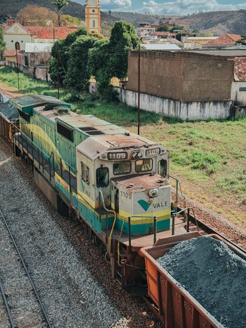 A vintage diesel locomotive marked with the number 1109 and the name VALE is seen on railway tracks during daylight, approaching a loading area next to a large open freight wagon filled with grey gravel or crushed stone. The locomotive is painted in a light blue, yellow, and cream colour scheme and is equipped with headlights, buffers, and various diagnostic panels. The surrounding environment includes green grassy areas and a small residential or industrial building in the background, with trees and hills under a clear sky. This scene illustrates the loading and transportation process of railway freight, which is relevant for logistics in house relocations and moving services as provided by Maida Vale Movers. The image captures the outdoor setting, highlighting the connection between transportation infrastructure and safe, efficient furniture and belongings transport during home removals.