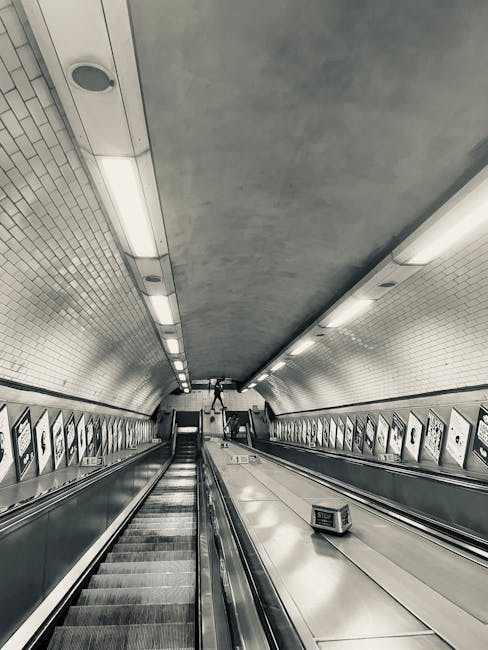 A black-and-white photograph of the underground station passageway showing a wide tunnel with a high, curved ceiling illuminated by long, rectangular fluorescent lights running parallel along the length of the passage. The walls are lined with small, square white tiles, and the floor features an automatic moving walkway with metallic handrails on either side. A person is visible at the far end of the walkway, pushing a trolley loaded with boxes wrapped in plastic and cardboard, indicating a home relocation or moving process. The scene suggests a transit point for transporting household items, with the person's luggage prepared for moving services offered by Maida Vale Movers, who may facilitate furniture transport and packing logistics as part of their removals services in the Maida Vale area.