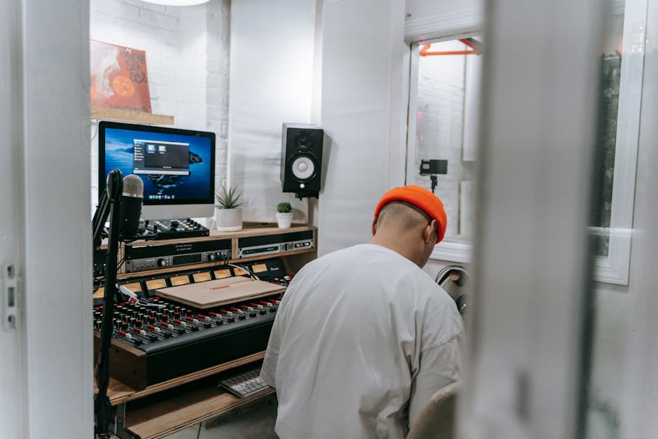 Inside a recording studio, a man wearing a plaid hat, headphones, and a blue jacket is seated at a large mixing console with numerous faders, knobs, and meters, engaged in home music production. In fr