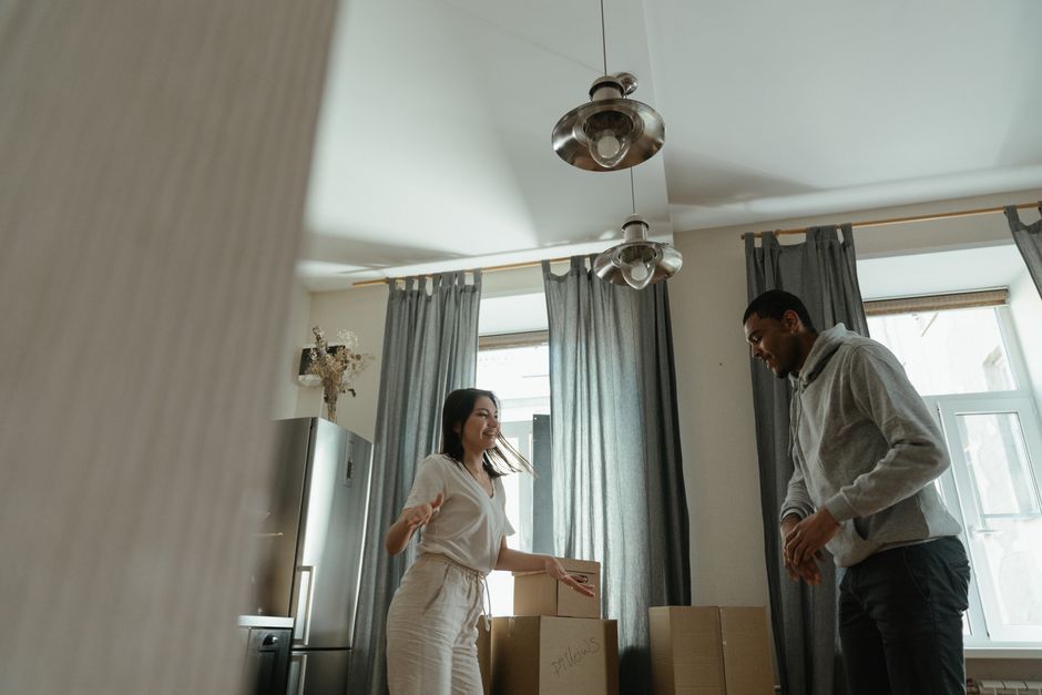 A man and a woman are engaged in home relocation activities outdoors. The woman, with long, curly hair pulled back in a bun, is standing beside an open hatchback car and is in the process of loading a