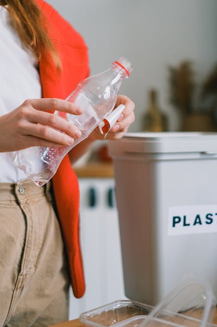 A house removal worker, dressed in an orange jumpsuit, stands inside a residential property holding two large blue plastic garbage bags filled with waste. The worker’s face is not visible, focusing 